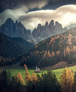 St Johann Church in the Dolomites in a green valley surrounded by evergreen forests, golden autumn trees, beyond are jagged rocky peaks under a cloudy sky.