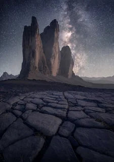 A dramatic night landscape photograph of Tre Cime, the three tall rock spires under a star-filled sky with the Milky Way, with cracked dark ground in the foreground.