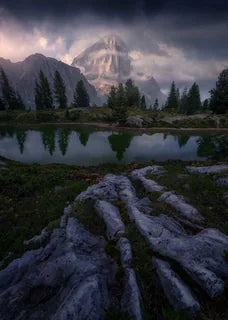 A dramatic mountain landscape in the Dolomites with a calm lake of Lago di Limides reflecting evergreen trees and a tall rocky peak, under a dark stormy sky. Foreground rocks lead the eye toward the reflective water and forested slopes while pink flowers cover the sides of the rocks.