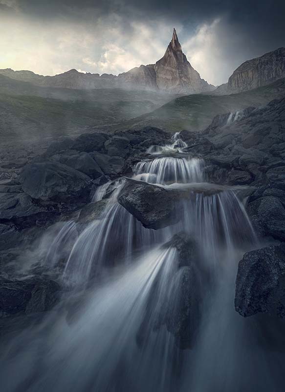 Aiguille Dibona in the French alps. A waterfall flows before the sharp mountain peak. Faint layers of mist cover the ground separating the peaks from the grassy hillside.