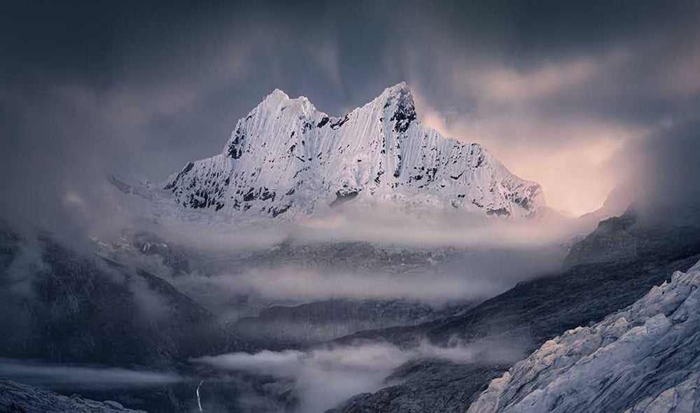 a giant white Andean mountain with snow and mist, backlit with a sunrise and stormy weather.