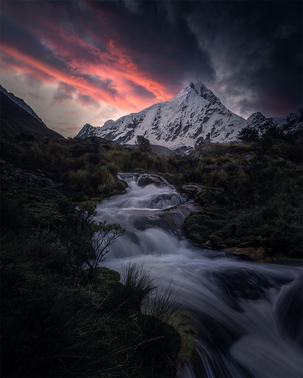 Copicalqui mountain at sunrise in Peru with a long exposure waterfall in the foreground, the giant snow covered peak underneath a vibrant stream of pink and blue clouds.