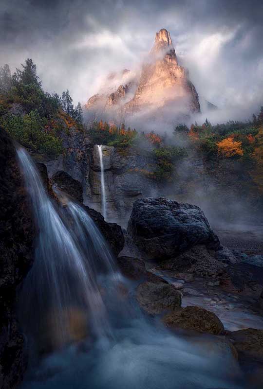 In the Dolomites a misty mountain towers above an autumn coloured ridgeline of trees. Below three waterfalls flow together into a stream. 