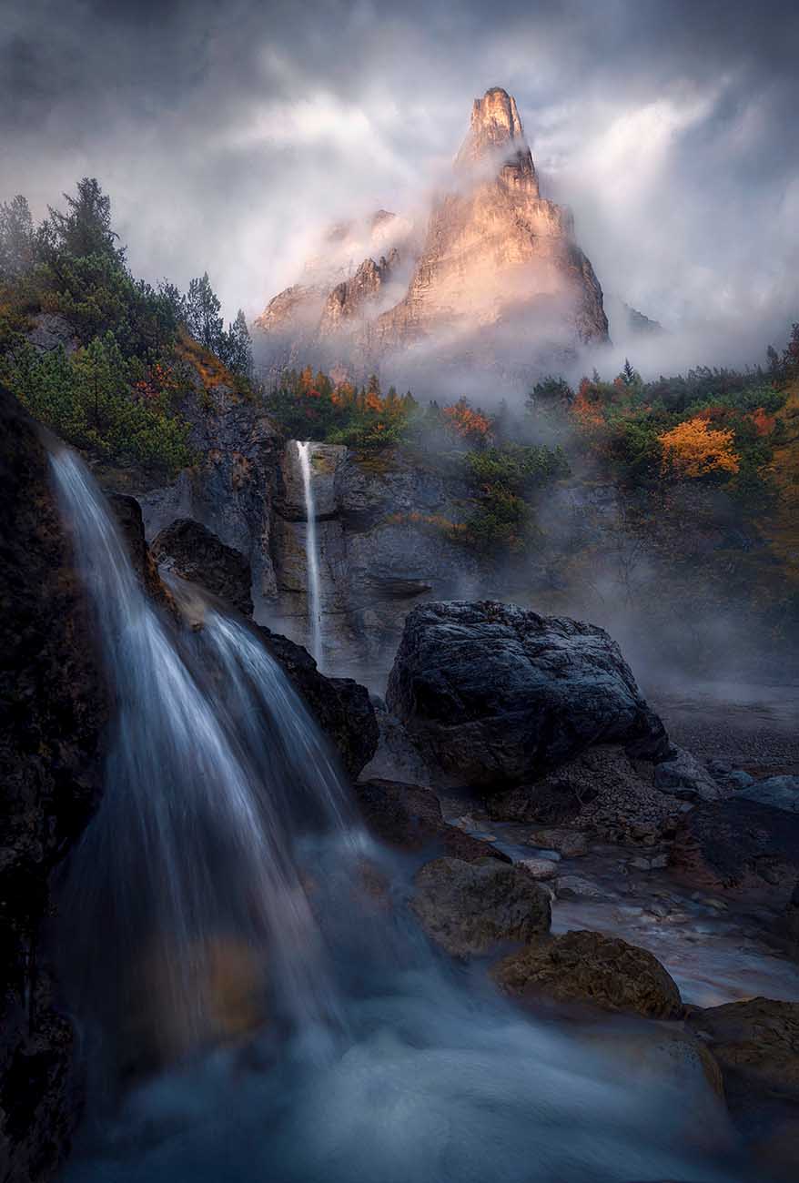 A tall castle shaped mountain peak in the dolomites surrounded by clouds and mist catching the last golden light, an autumn coloured forest beneath the peaks with two tall waterfalls in the foreground.