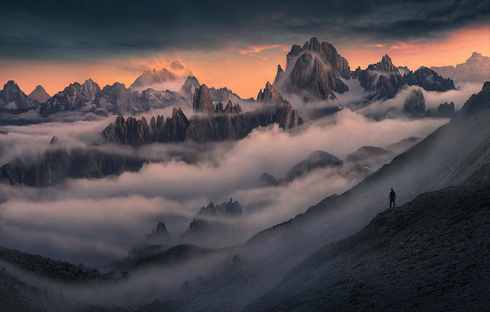 A person stands on the edge of a ridgeline looking out at layers of mountain peaks shrouded with layers of mist in the dolomites at sunset, the sky is a firey pink and orange glow.