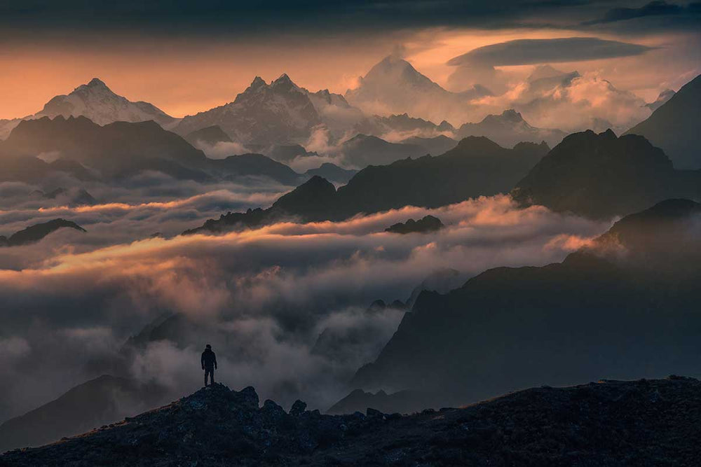 A silhouette of a man standing on the ridge of a high rocky mountain in the Himalayas of Nepal. He looks out at the pink sunset, layers of clouds and peaks including Mount Everest.