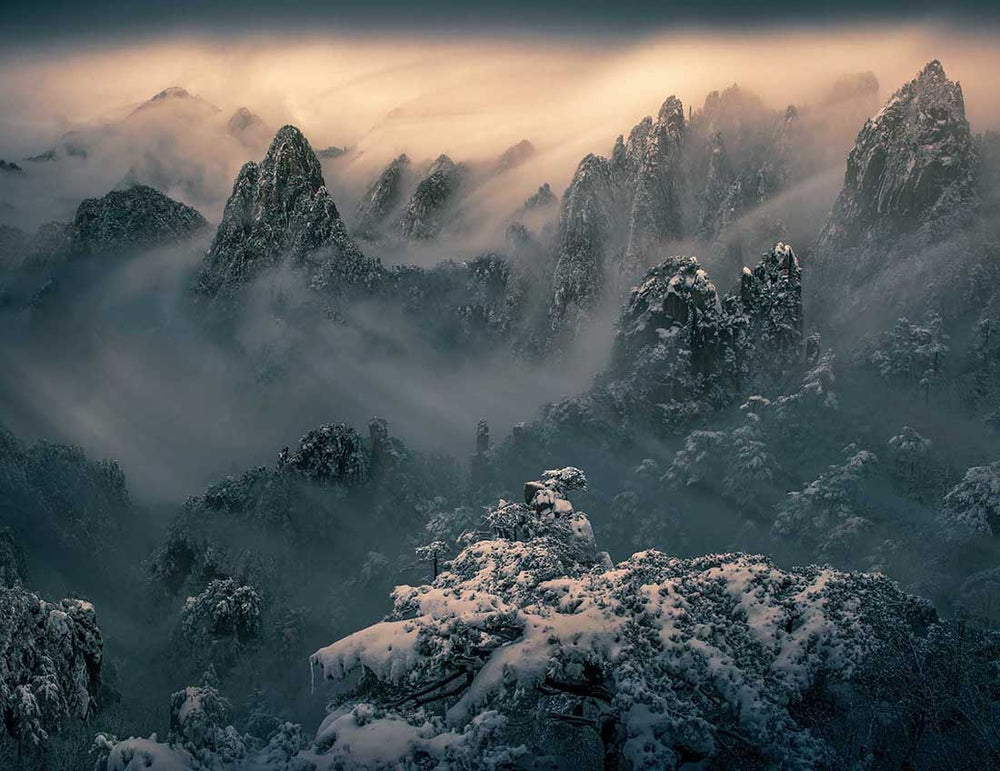 Huangshan mountain in winter high above the snow covered trees. The orange sunrise light is obscured by the thick clouds and mist rolling through the sharp rock faces.