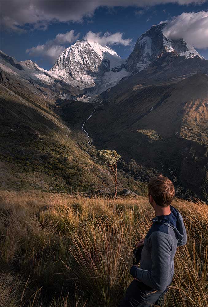A person stands in front of the Huascaran mountains in Peru during the afternoon sunlight. A long tree stands in the midground in front of a waterfall.