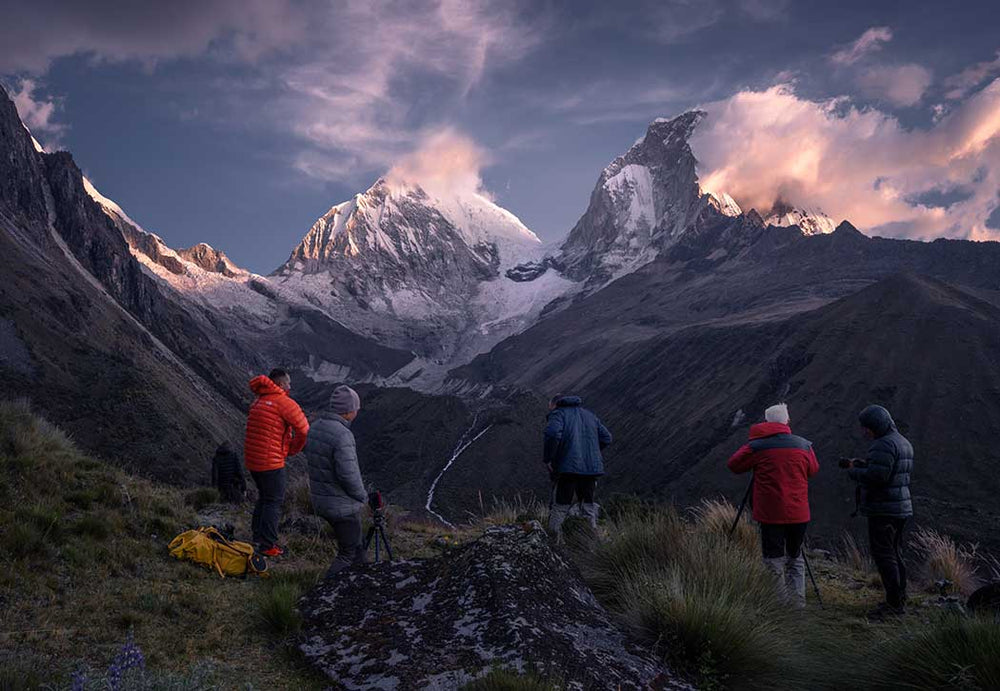 A group of people stand before the snow covered Huascaran mountain at sunrise. The clouds are pink in the morning light above the peaks.