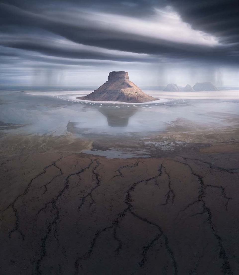 A dramatic image of salt flats in kazakhstan with a large isolated rock formation in the middle of the mudflat and reflected in the shine of water, in the distance are rain showers and some more peaks, in the foreground dried mud creates a network of river patterns, a dark moody sky is above.