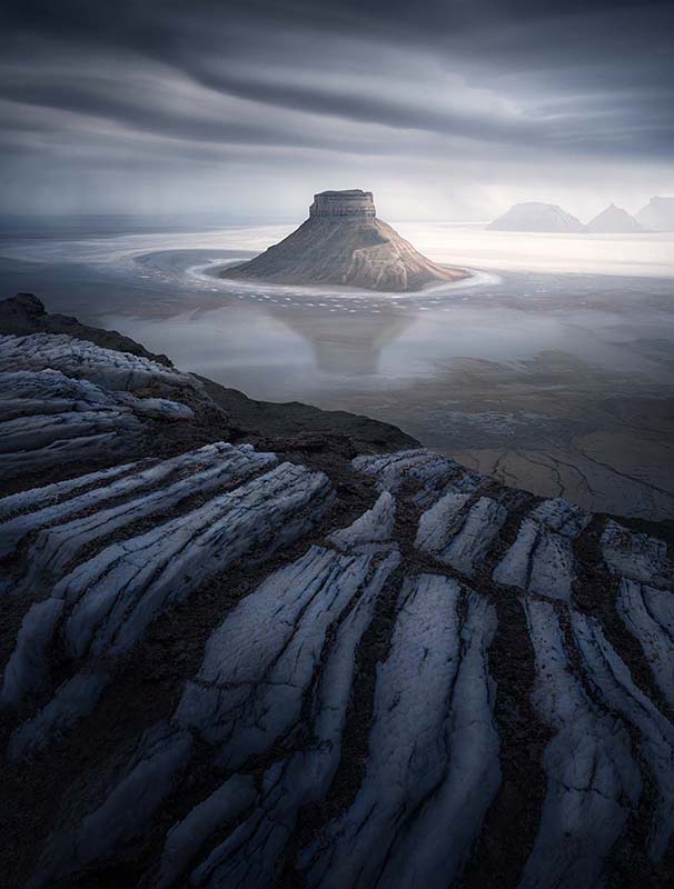 Kazakhstan salt flats show a lonely mountain in the middle of what used to be the earths sea bed. Lines of rock formations leads towards the mountain. It's reflection shines in the recently rained upon salty mud.