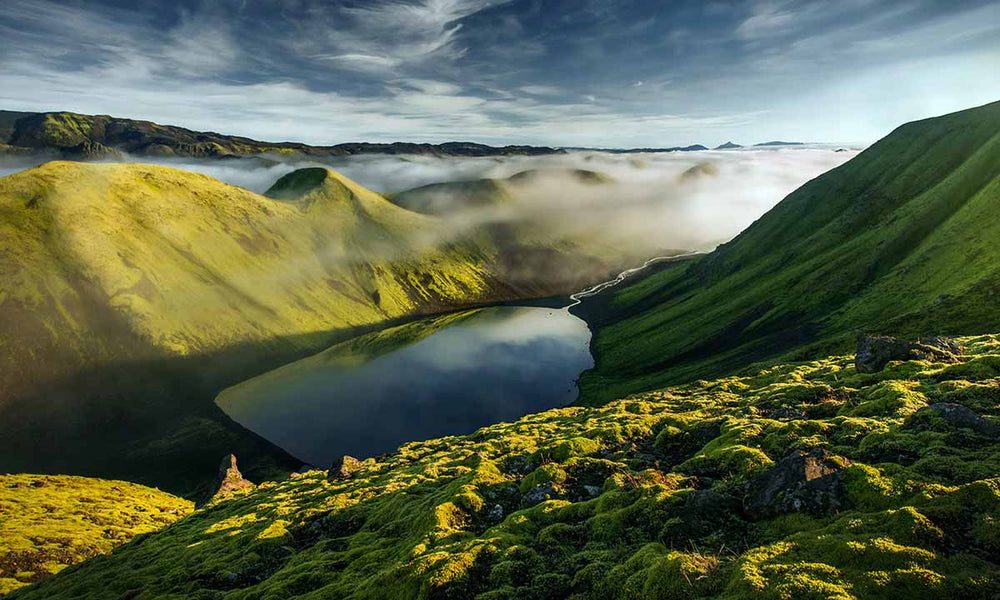 Langisjor in Iceland at Sunrise. A cloud inversion covers the base of the mountains in the valley leaving just the peaks and mounds showing. In the foreground is a lake with a stream leading into the distance.