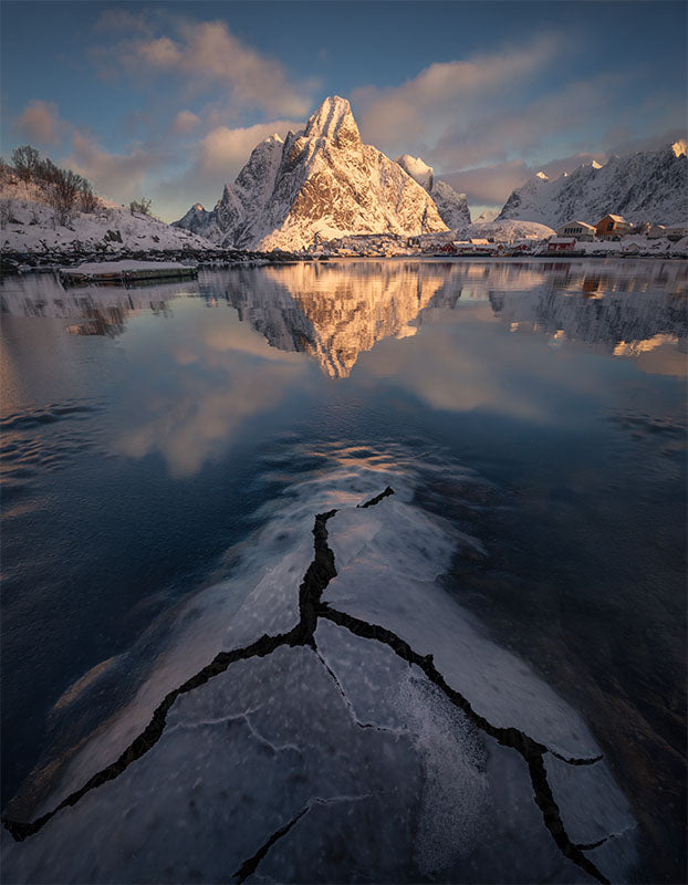 Sunrise at olstinden in Lofoten is lit up with golden light and pink clouds. in the foreground is a giant domed icecrack leading into the reflective lake.