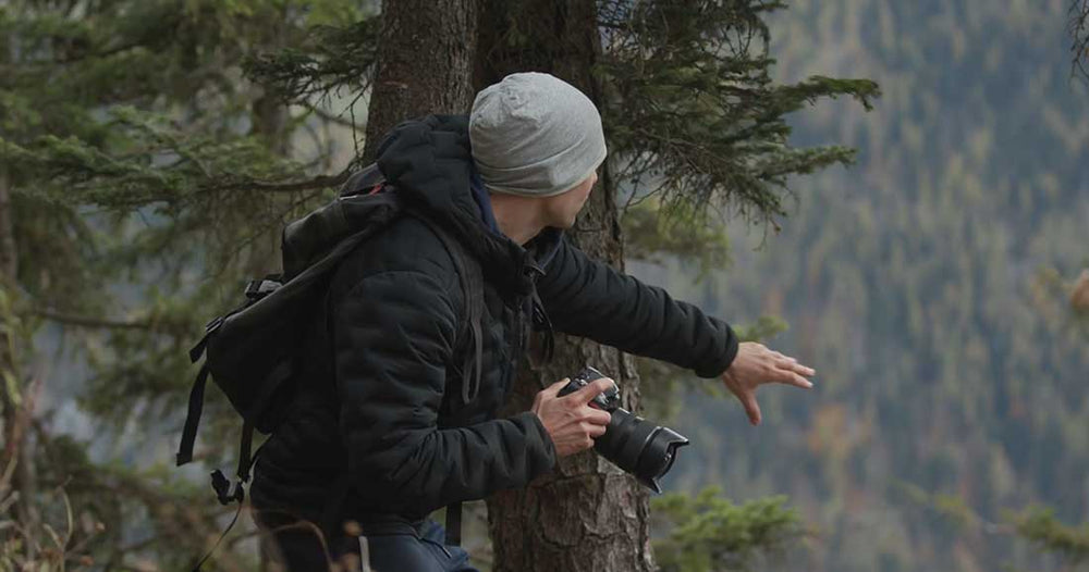 A man with a camera searches for compositions in a forest wearing a beanie and backpack.