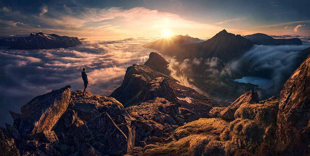 A man stands on the ridgeline of a high fjord in northern Norway during sunrise. A cloud inversion can be seen in the golden light between the mountains. An ultra blue lake fjord is visible to the right of the sun.