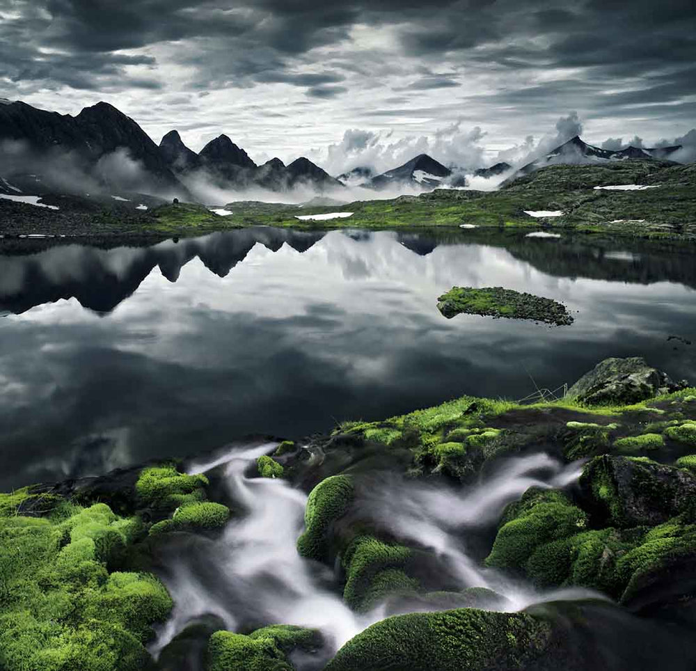 A mist covered mountain range is reflected in a glassy lake which is surrounded by waterfalls and green moss.
