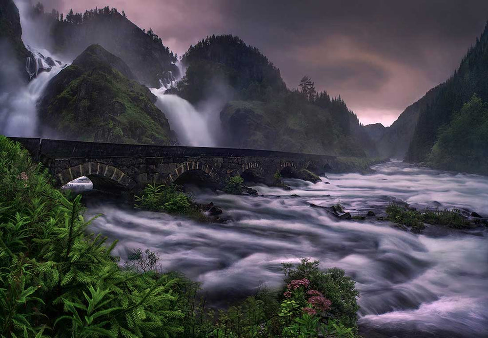 Latefossen Waterfall in Norway during a pink sunset. the waterfall flows down the mountain and underneath a bridge with pretty pink flowers adorning the river edge.