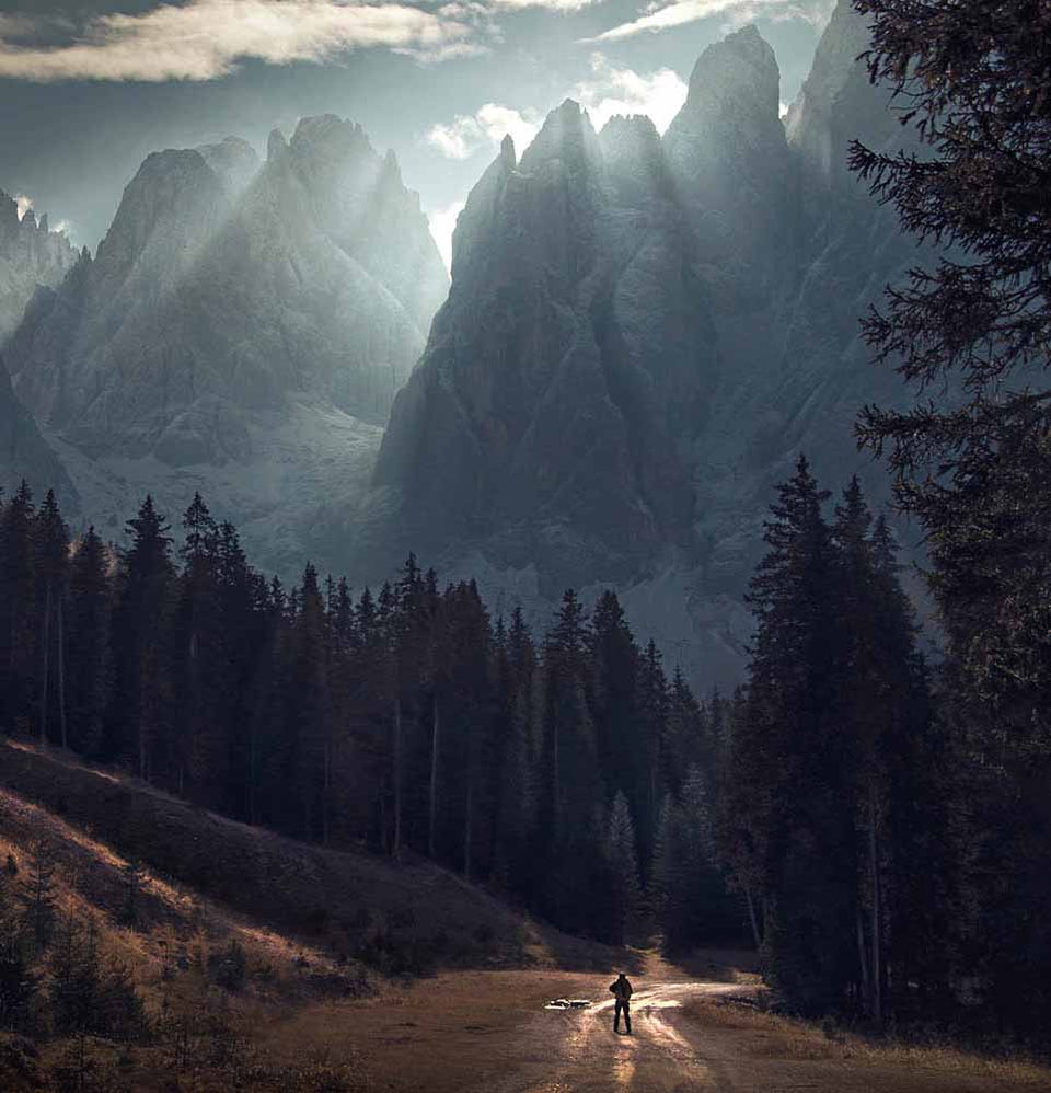A dramatic mountain landscape with jagged rocky peaks, sun rays breaking through clouds, a dark evergreen forest, and a small silhouetted figure standing on a dirt road in the foreground.