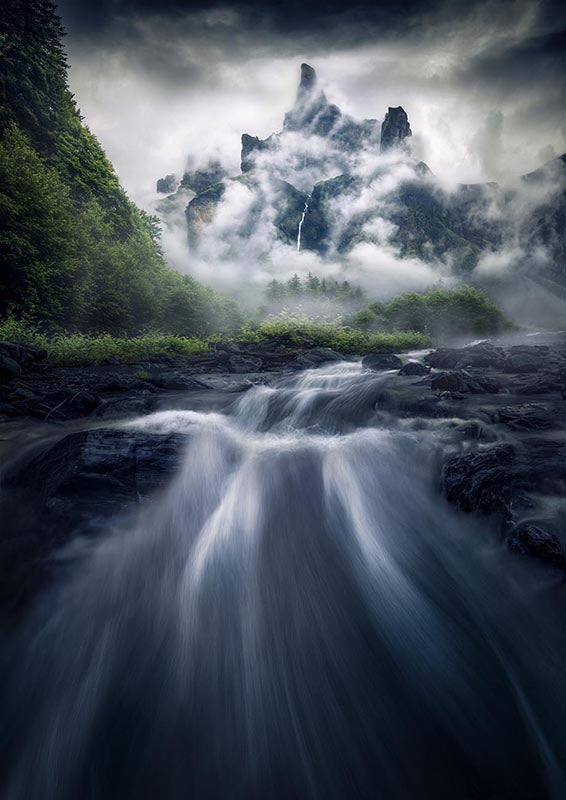 Sixt-Fer-a-Cheval a stream flows through a misty forest to a misty mountain with waterfalls running down the sides.