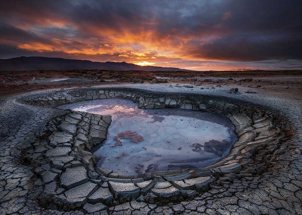 Theistareykir in Iceland at sunset. The sun glows in orange and purple on the horizon colouring the clouds above it. In the foreground is a shallow lake embedded within the giant mud cracks surrounding it.