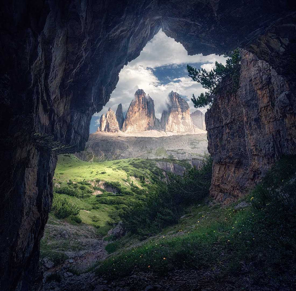 Drei Zinnen in summertime as seen from a cave-view, in the midground a grassy meadow of flowers and trees make the hillside vibrant.