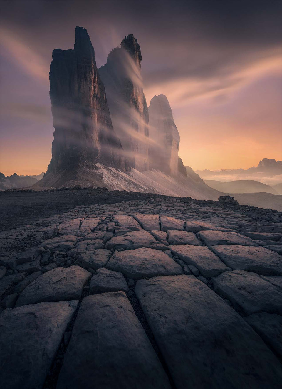 A dramatic landscape photograph of three towering rock spires in the Dolomites, with mist swirling around the peaks and a cracked stone foreground under a warm orange and purple sky.