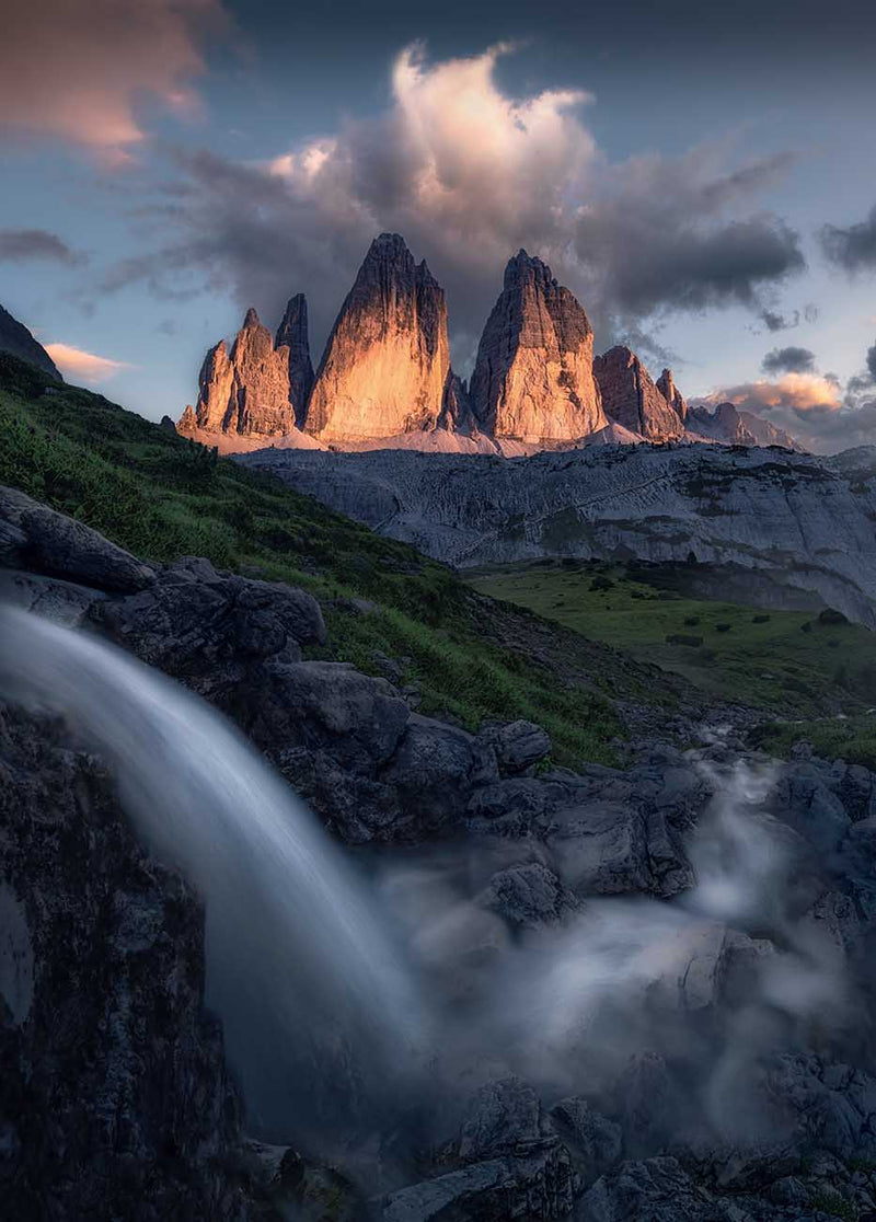 A dramatic mountain landscape of Drei Zinnen, a large waterfall in the foreground from the left flows right and towards the peaks, green slopes, and clouds lit by warm light near the peaks.