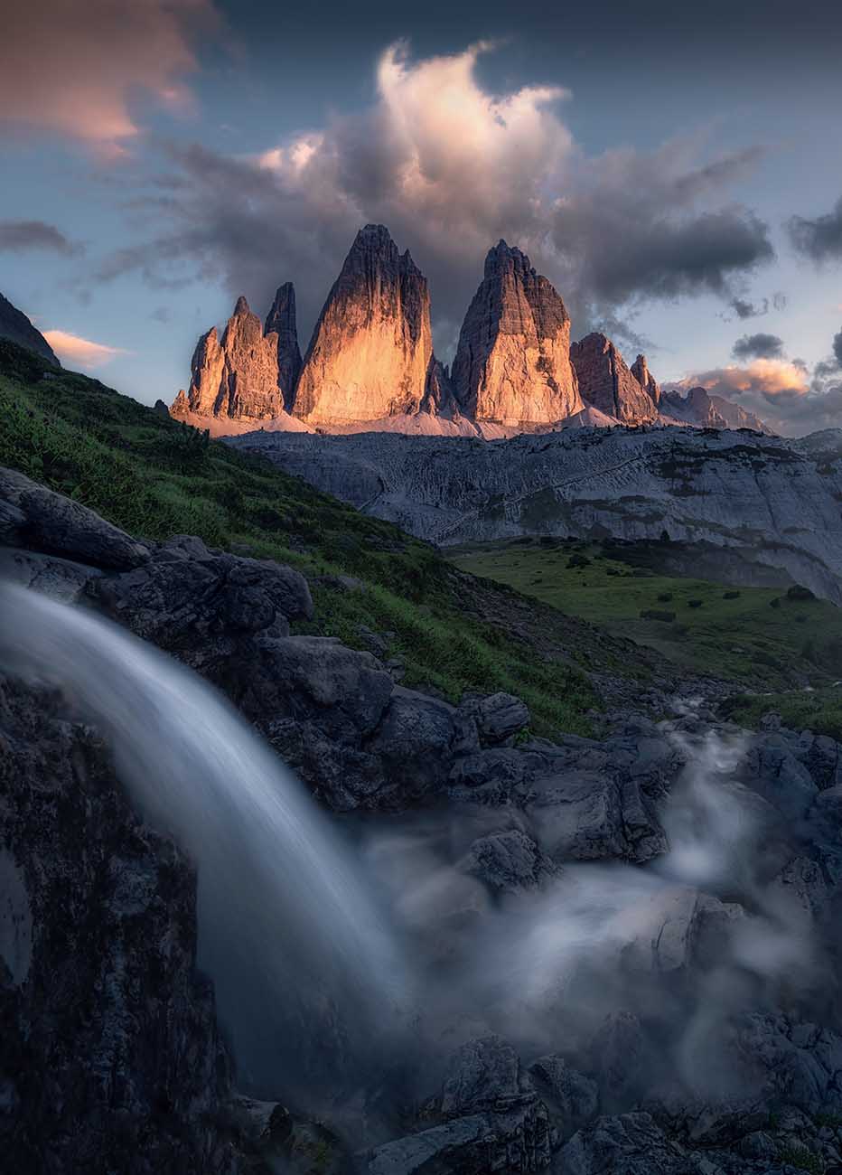 A dramatic mountain landscape of Drei Zinnen, a large waterfall in the foreground from the left flows right and towards the peaks, green slopes, and clouds lit by warm light near the peaks.