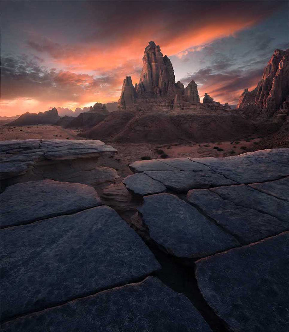 A dramatic desert landscape featuring tall rock spires under a fiery orange and dark blue sunset sky, with textured stone pavement in the foreground.
