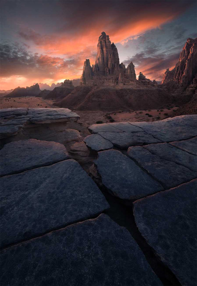 Cracks in a broad rock surface leading toward a sandstone tower beneath a red sky in Saudi Arabia.