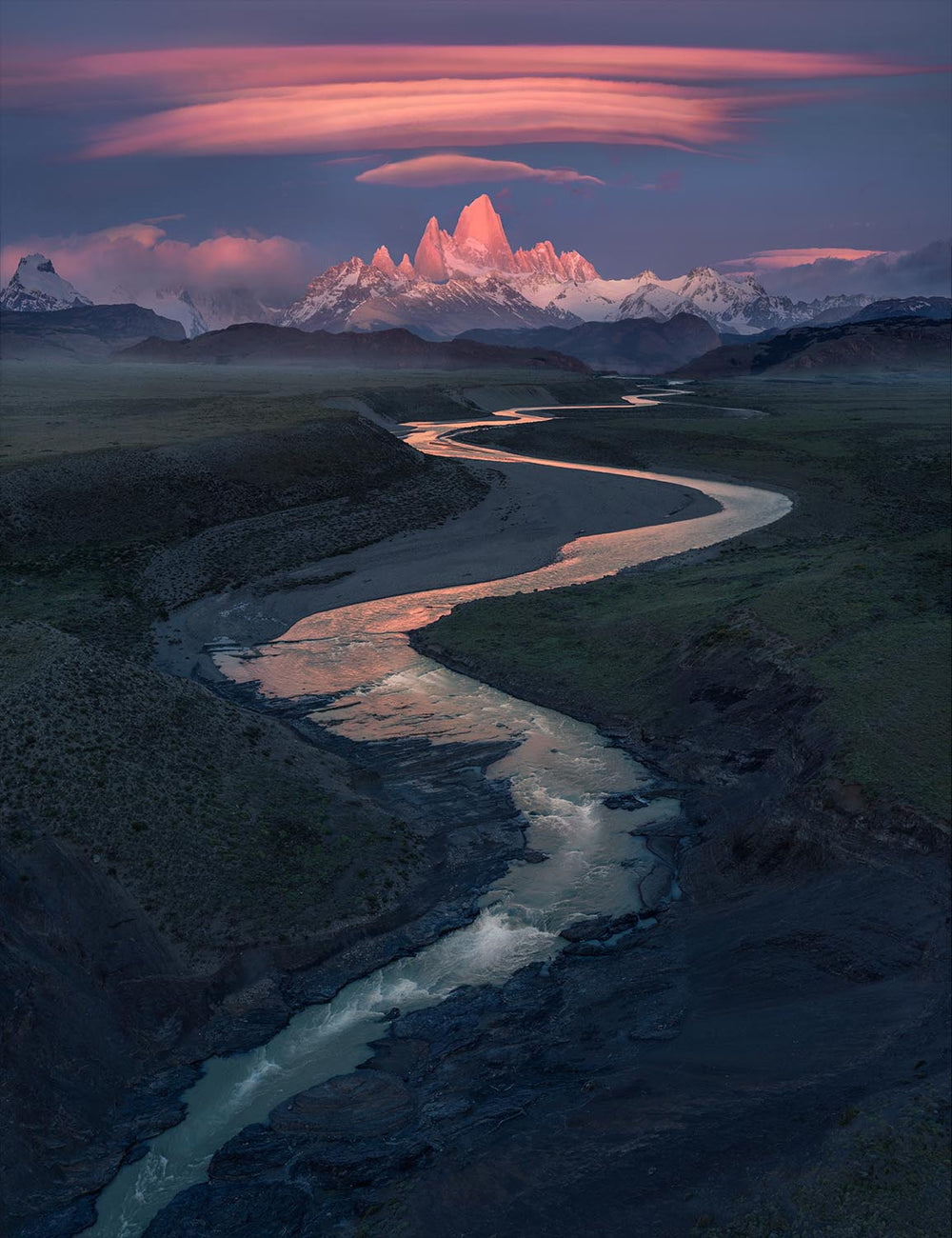 ariel view of monte fitzroy under a pink sunrise taken by drone. A winding river leads towards the mountain in Patagonia.