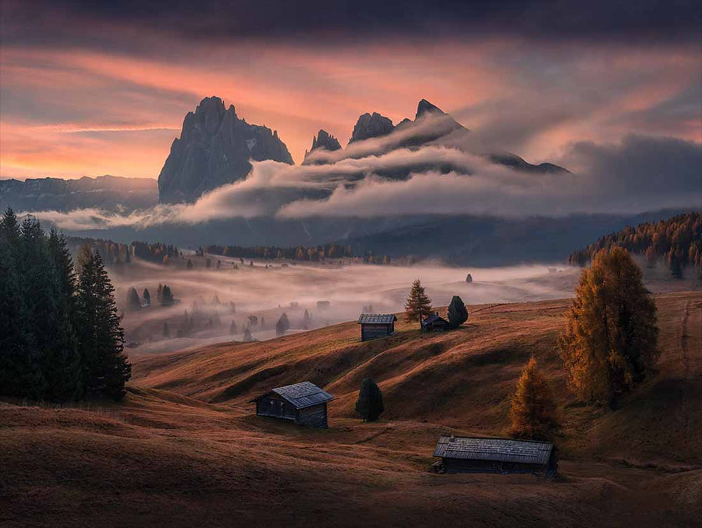 Alpe di siusi mountains on an autumn morning sunrise. The clouds are pink and mist rolls through the trees below. Fall coloured trees frame the scene.