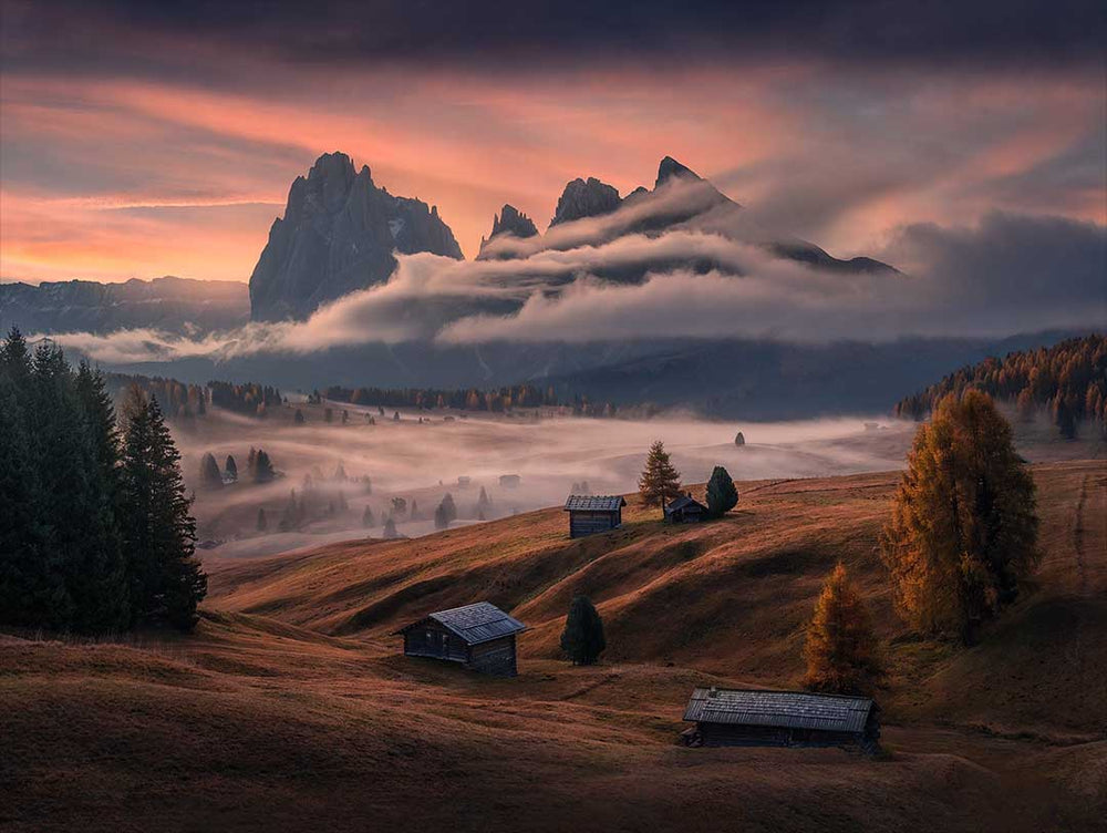 Alpe di Siusi in autumn at sunrise, mist streams along the peaks and through the forest alpine meadow. Mountain huts scatter the hills and a pink firey sky lights up the clouds.