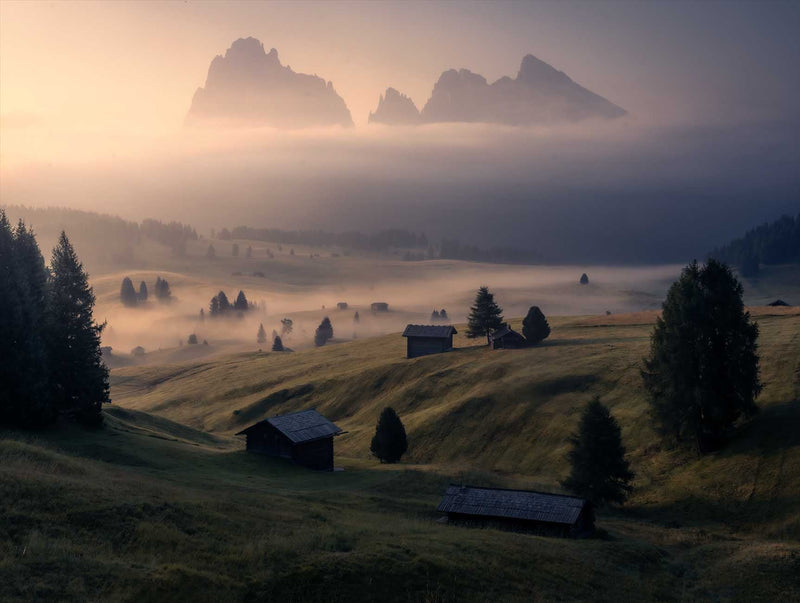 A misty mountain valley landscape at sunrise, with rolling grassy hills, scattered dark trees, small rustic cabins, and jagged mountain peaks fading into low fog.