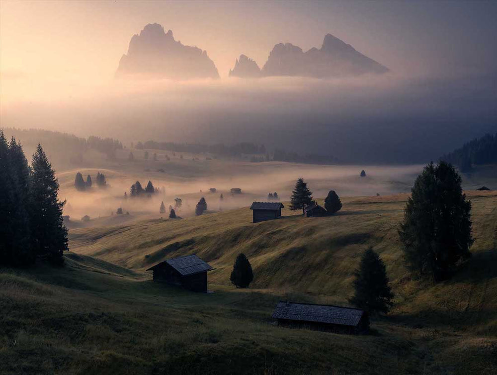 Alpe di Siusse hillside with mountain huts and mist. The warm sunrise glow illuminates the mountain peaks.