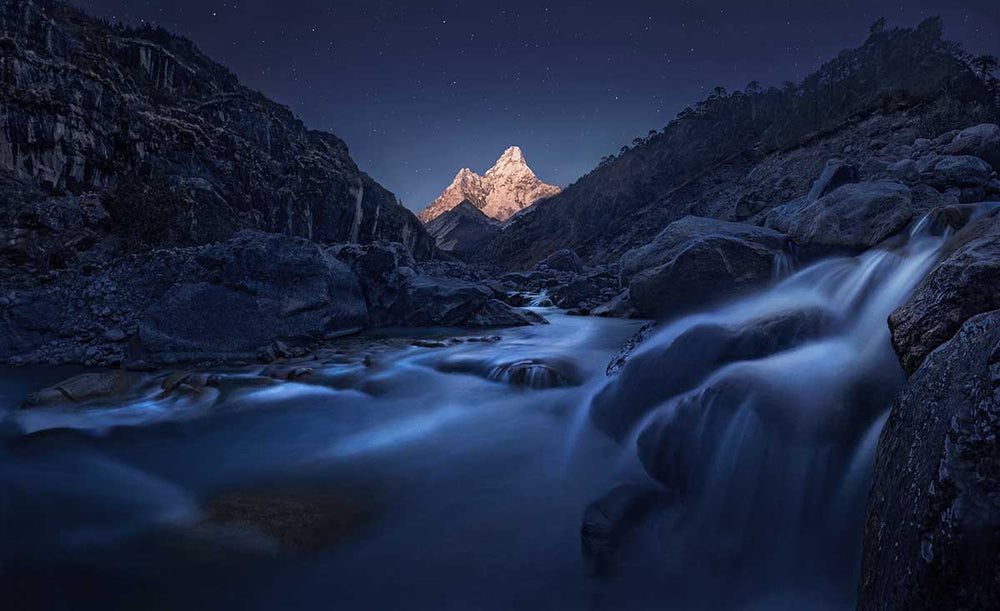 Ama Dablam in the Himalayas of Nepal during the night, The peaks of the mountain glow in the moonlight. in the foreground a stream and waterfall runs through the valley to the mountain.