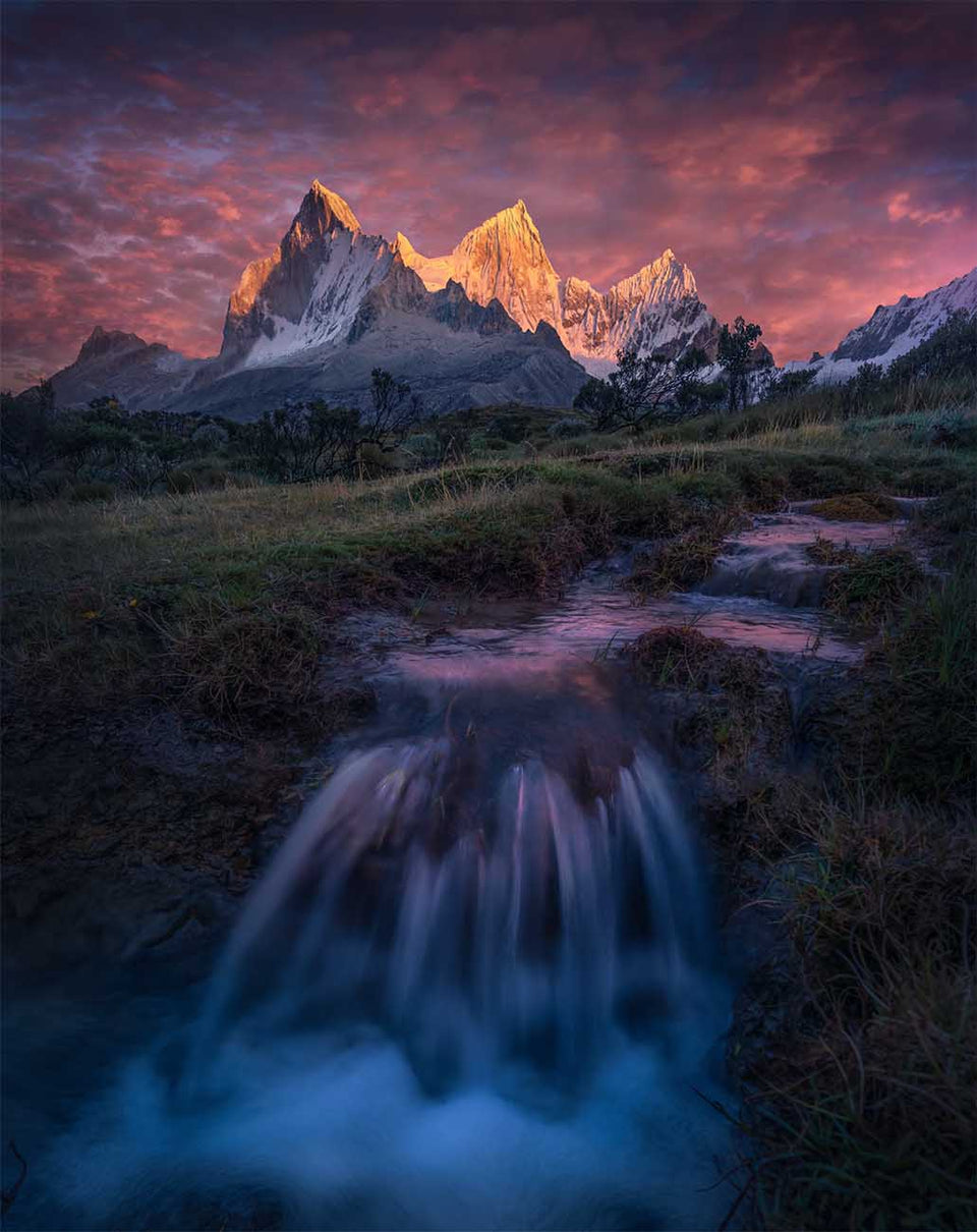 Snow-covered Peruvian mountains glowing in the evening above a meadow and a small waterfall.