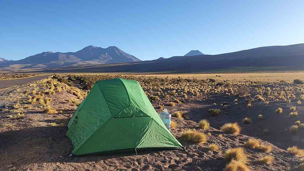 a green tent pitched by the side of a highway in the Atacama desert, volcanos can be seen in the background.