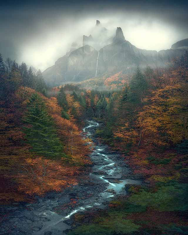 Autumn coloured trees line the valley beneath a mountain in France. A stream winds its way through the orange coloured valley. Behind a misty mountain stands tall, from it flows a waterfall.