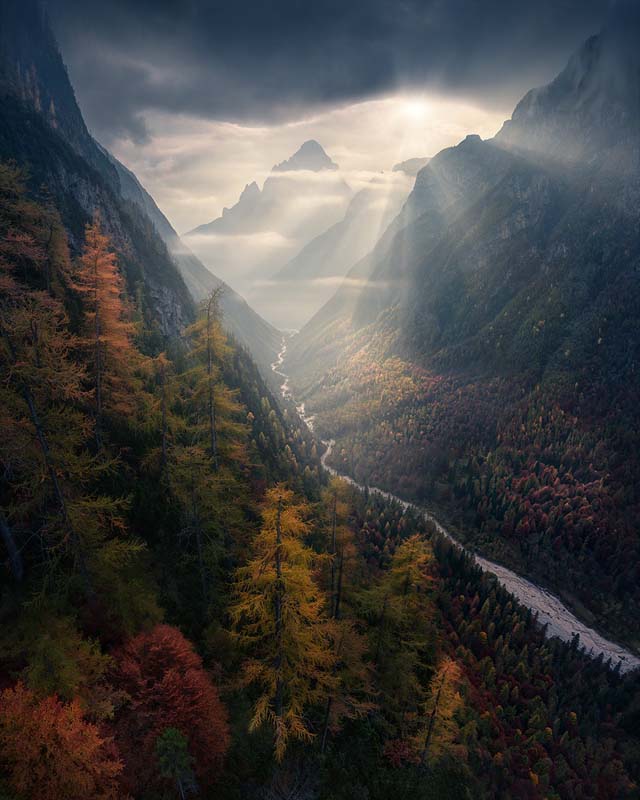 Sunrays at sunrise in the Dolomites flow into an autumn coloured valley of trees and winding river to the mountains beyond. Taken by Max Rive.