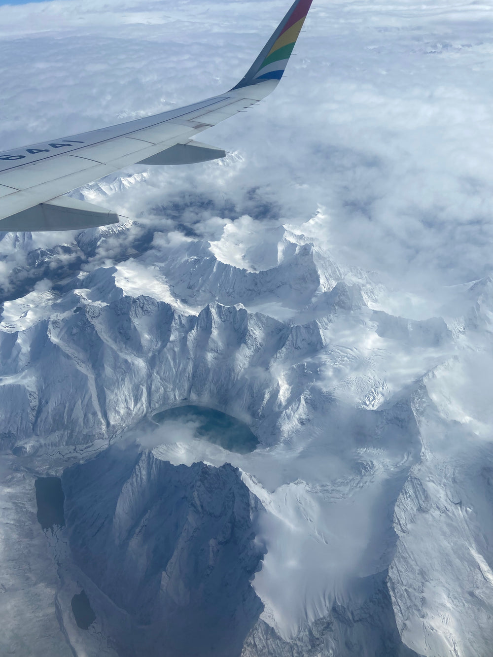 The view out of the window of a plane where the wing is visible, beneath is a sea of snow covered mountain peaks and a high mountain glacier lake.