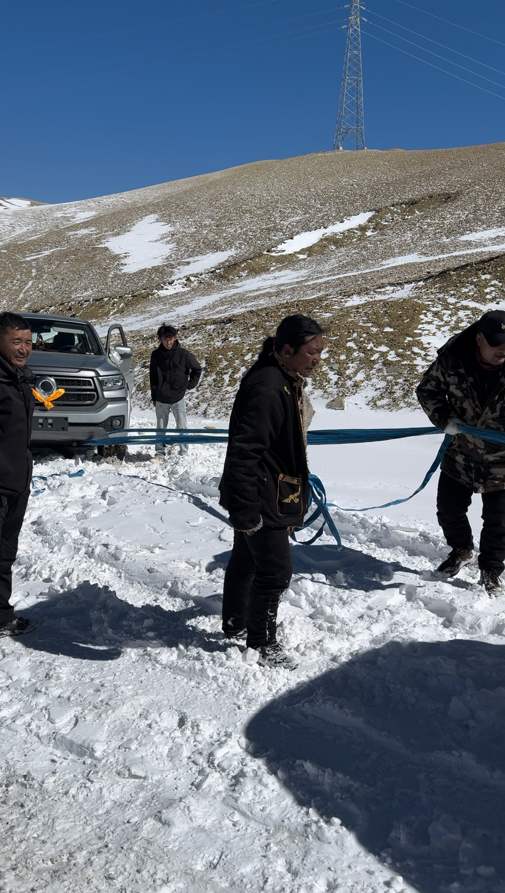 A snowy scene with a car stuck in snow beside a road in the mountains, four people are busy attaching a rope and chains to pull the car out of the snow.