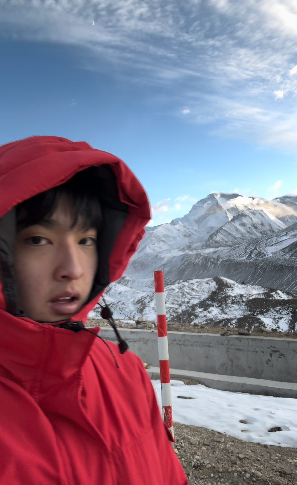 A selfie of Forest Kai in a red jacket in the high mountains of the Himalayas in Tibet, he looks cold surrounded by snowy mountain peaks.