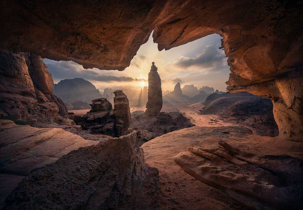 A cave in Saudi Arabia shows the sunrise light over the desert rocks. A large mountain in the middle is the subject and appears like a rocket ship formed by sandstone.