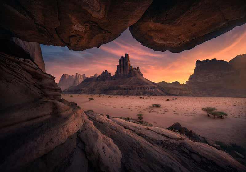A sandstone peak beneath a pink sky, framed by an overhanging cave in Saudi Arabia.