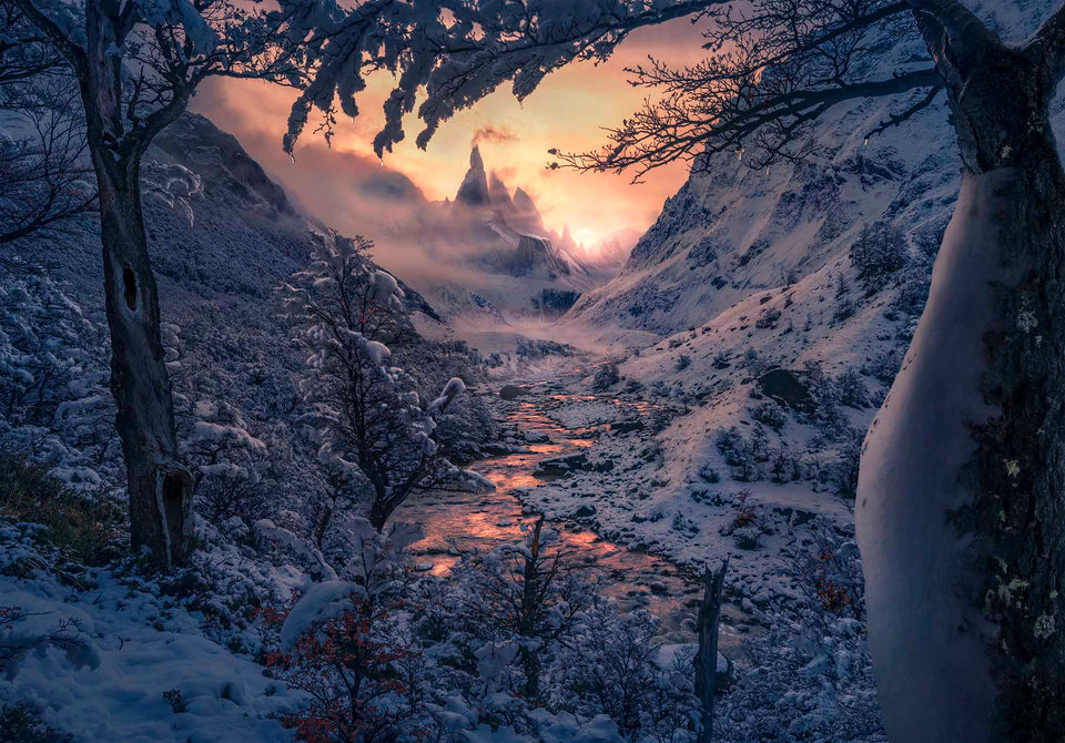 A snow covered mountain valley at sunset, with snow-covered trees framing a winding stream and misty peaks of Cerro Torre glowing orange in the distance.
