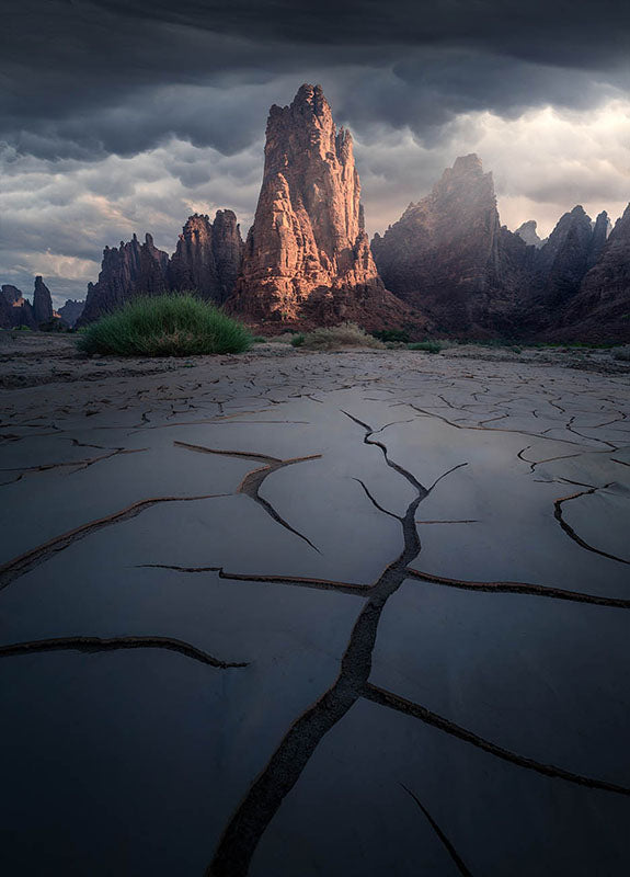 Giant mud cracks in Saudi Arabia beneath the sandstone rock formations. sunset light hits the side of the mountain and create soft light. layers of mountain can be seen either side.