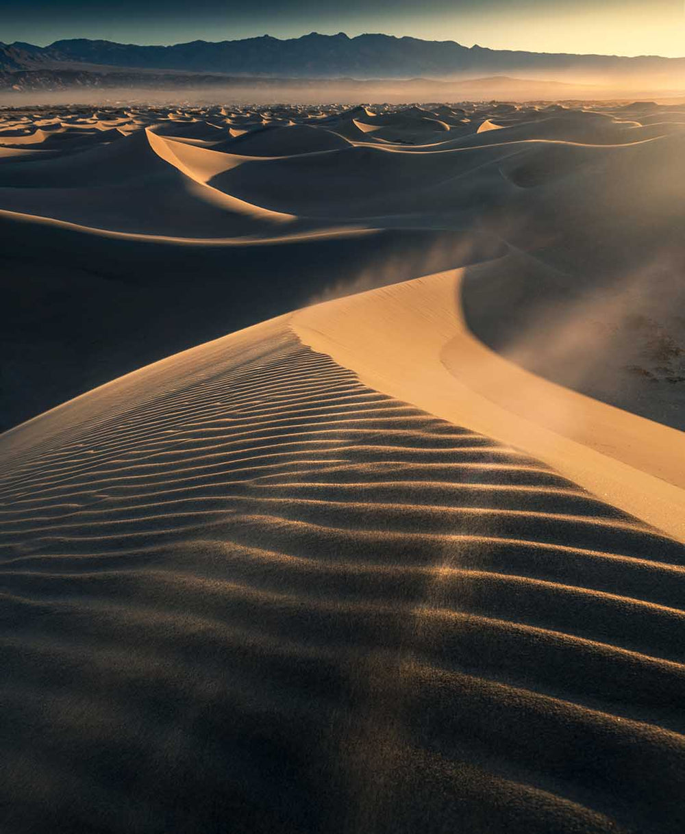 A desert storm in death valley creates streams of sand blowing across the layers of dunes into the golden sunset. Across the ridgeline of the main dune is lines and rivers of sand which have been shaped by time.