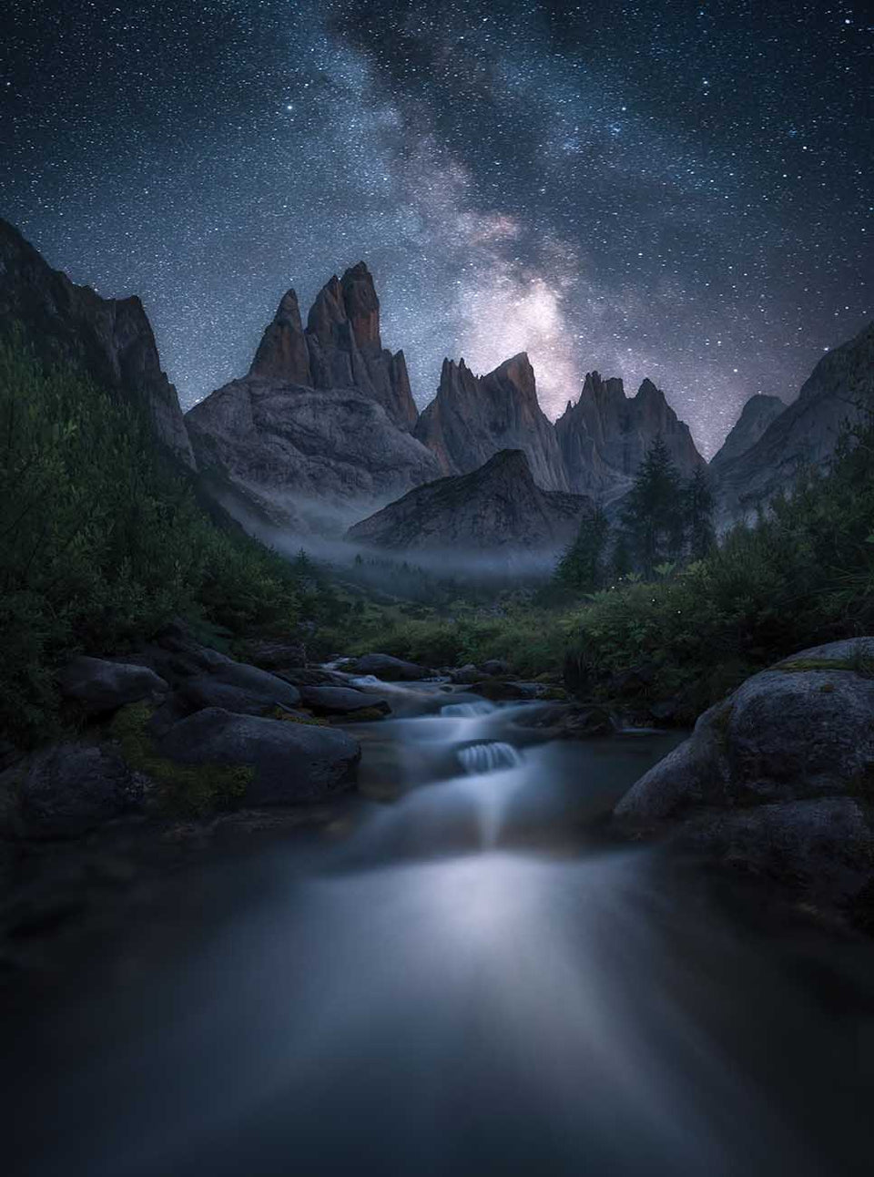 A mountain landscape in the dolomites with a stream flowing towards the mountains through a valley, stars and the milky way light up the sky, a forested valley is misty beneath the peaks.