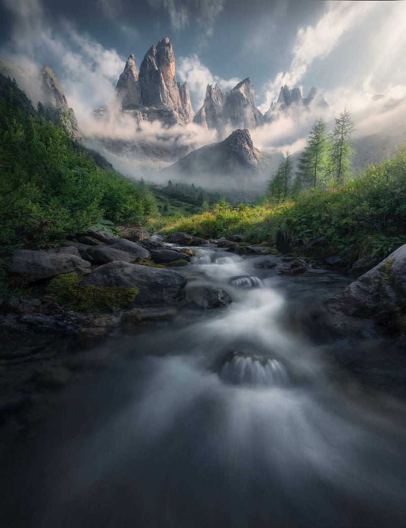 A dramatic mountain landscape with jagged rock peaks rising through mist and clouds, a flowing stream in the foreground, and green forested slopes under a cloudy sky.